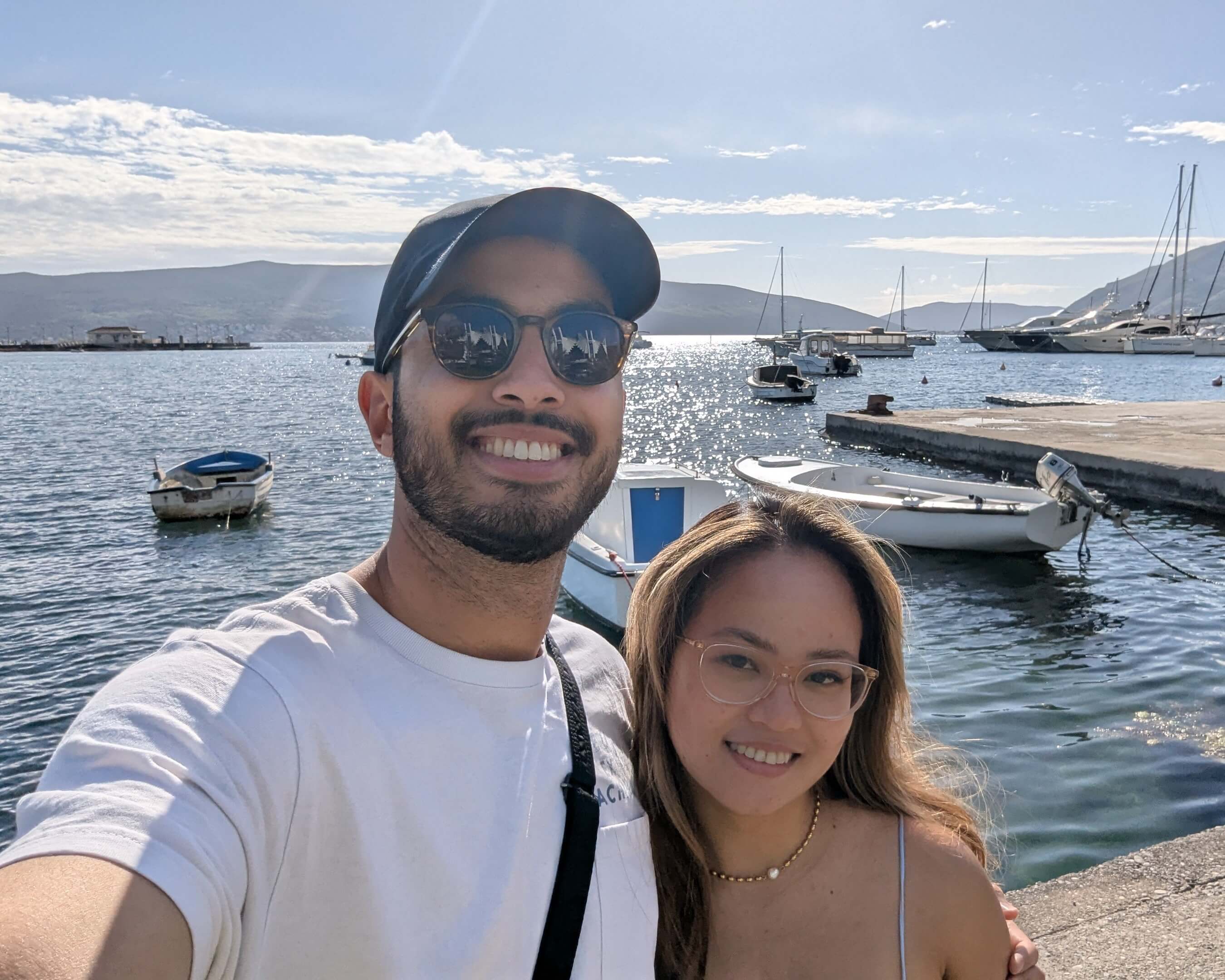 Two people taking a selfie by a waterfront with boats and mountains in the background.
