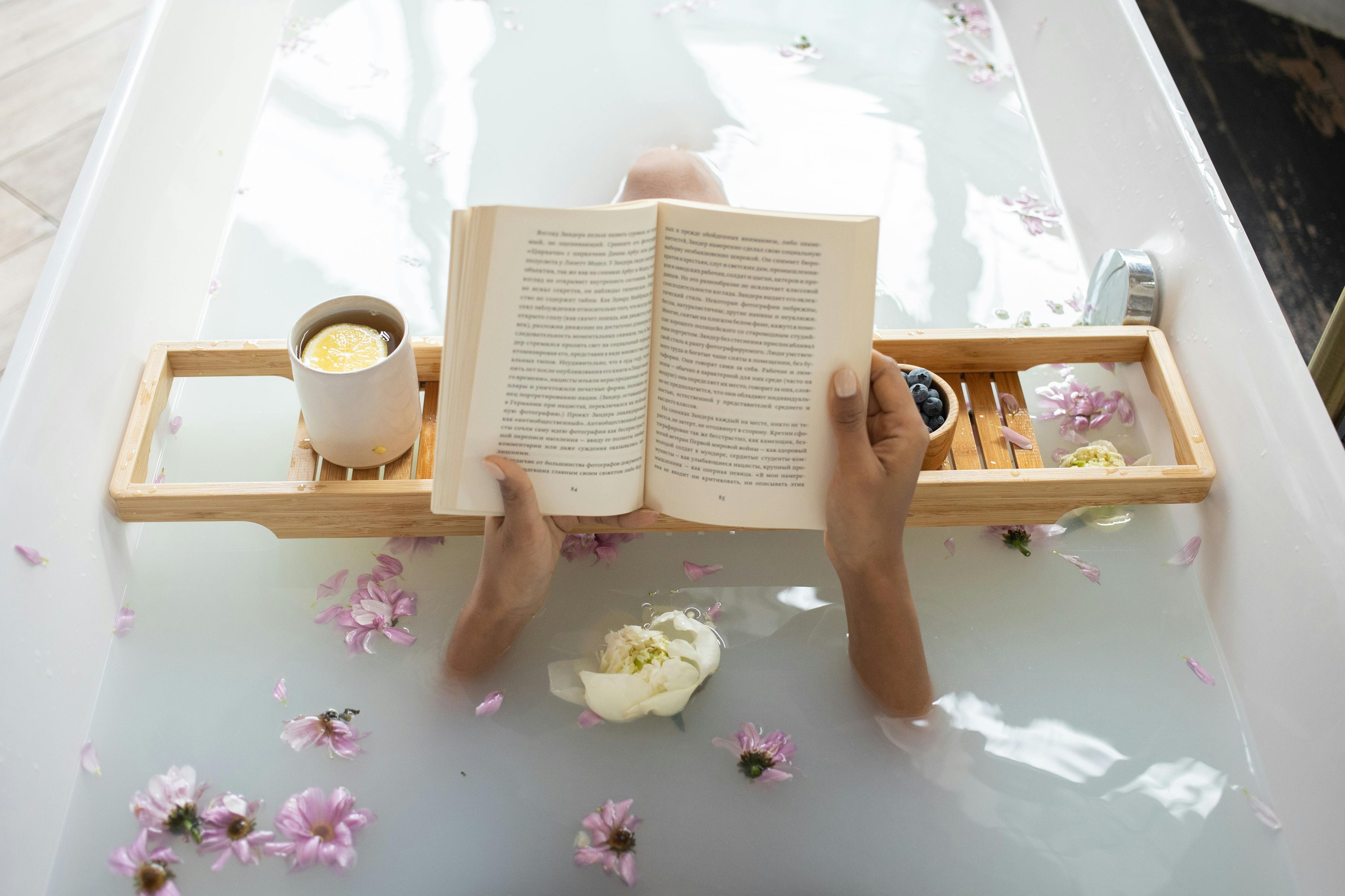 Person sitting in a bathtub, reading a book on a tray with a cup and flowers on a light surface.