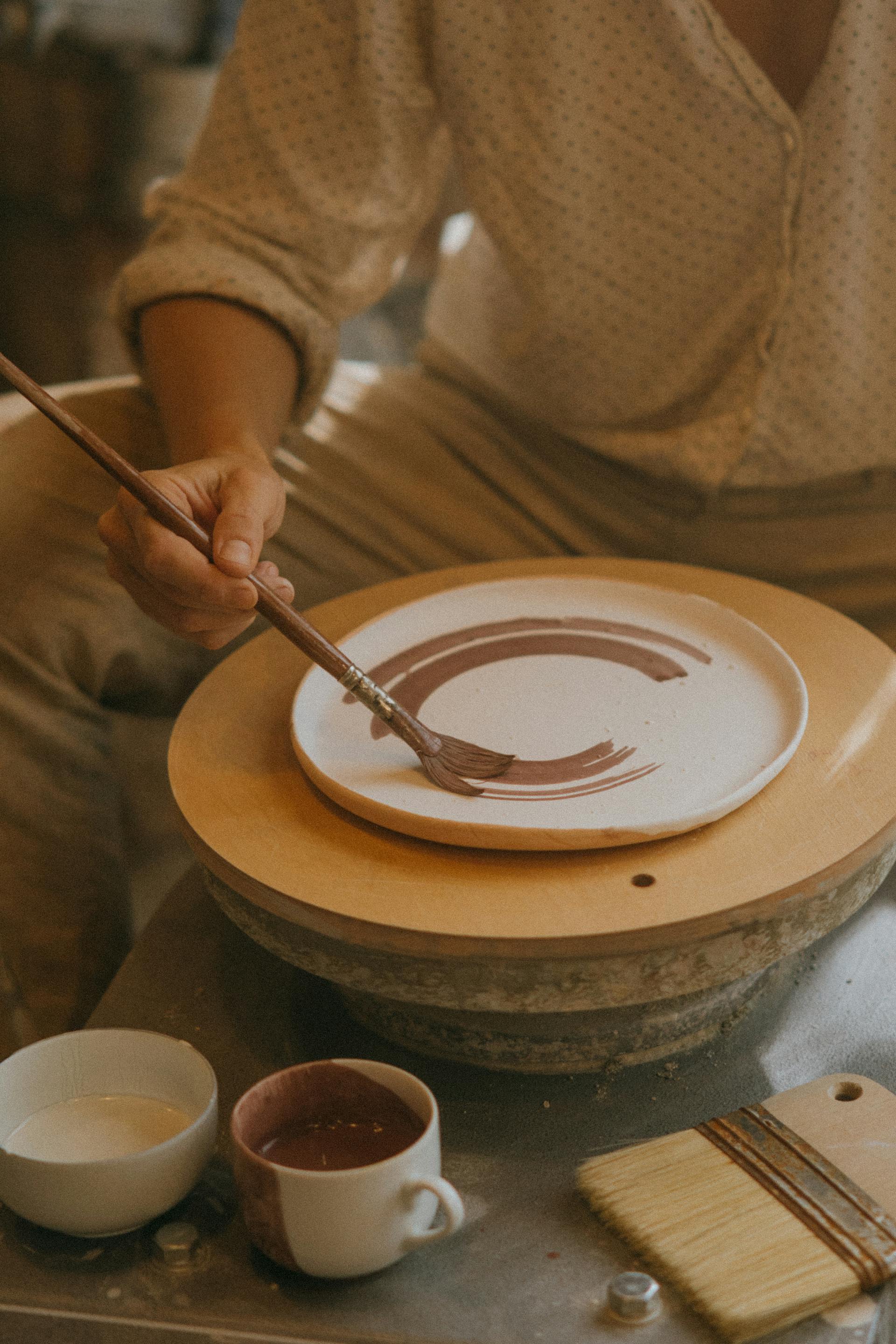 Person painting a ceramic plate on a potter's wheel with various pottery tools and materials around.